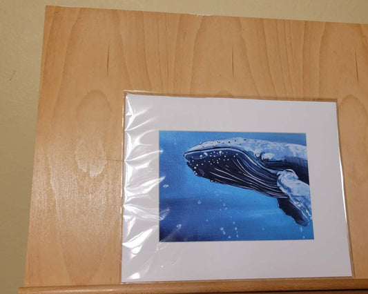 A framed whale art print with a white mat, wrapped in protective plastic, displayed on a light wood easel against a beige wall.